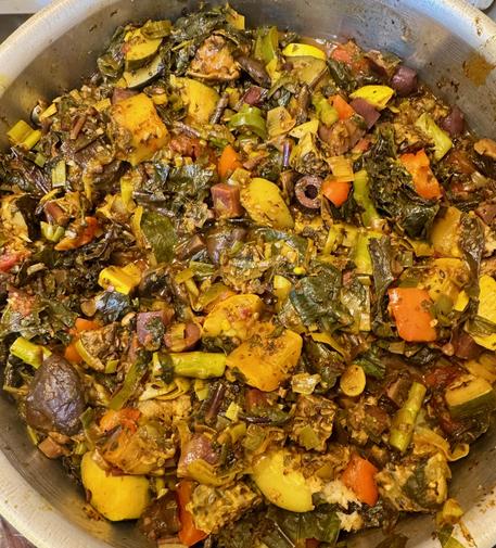 A big pot of stew showing yellow summer squash, diced purple potato, sliced black olives, greens, red bell pepper, and leeks, with an overall orange hue from the turmeric.