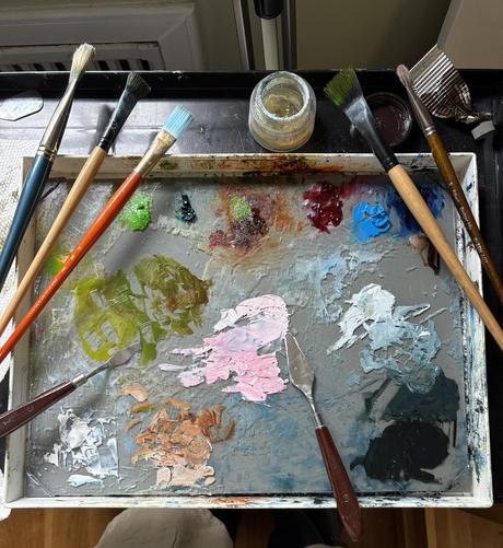 Photo of my paint palette today. There are puddles of oil paint on the gray glass palette, with an array of brushes and palette knives resting on the edges of the palette box.