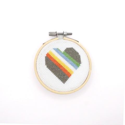 A photograph of a cross-stitch rendering of the Disability pride flag as a heart. It's in a light blonde hoop and on white cloth. The flag itself is a muted dark gray, red, yellow, white, light blue, and green.