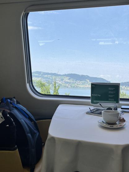 Blick aus einem Zug: Kaffeetasse, Bordrestaurant drin, Sonne und See vor dem Fenster.