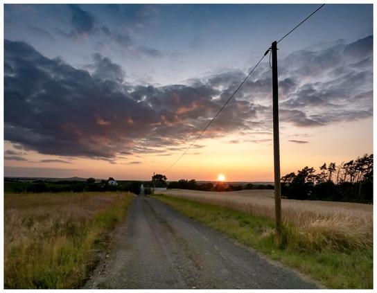 A gravel road leads through a field at sunset, with the sun dipping below the horizon. The sky is filled with colorful clouds, and a utility pole stands alongside the road. In the distance, a farmhouse is visible among trees.