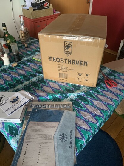 Some frosthaven stuff in front of the box with the frosthaven box in it, on a table