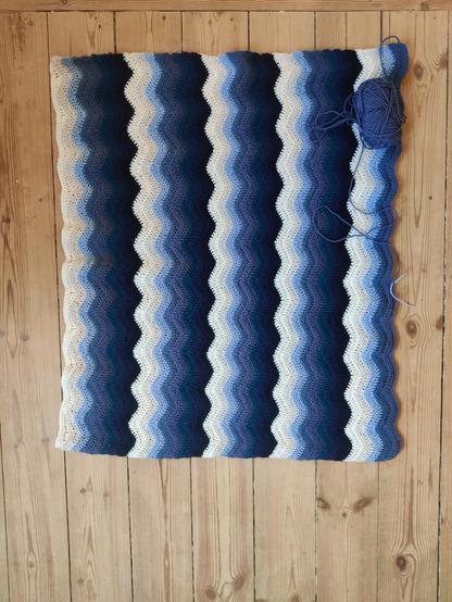 A crocheted blanket lying on the floor. It has a ripple pattern that looks kind of like waves in the sea, with each wave having a gradient of colour from dark blue to white 