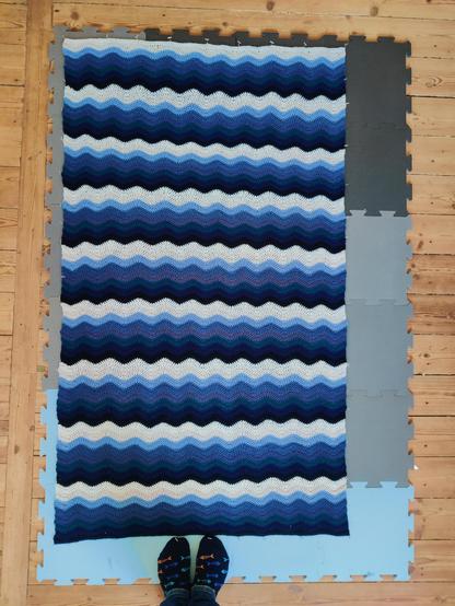 A ripple stitch crochet banket, laid out on foam squares for blocking. The blanket consists of nine repeats of shades of blue, from pale to dark, sort of like waves in the ocean seen from above 