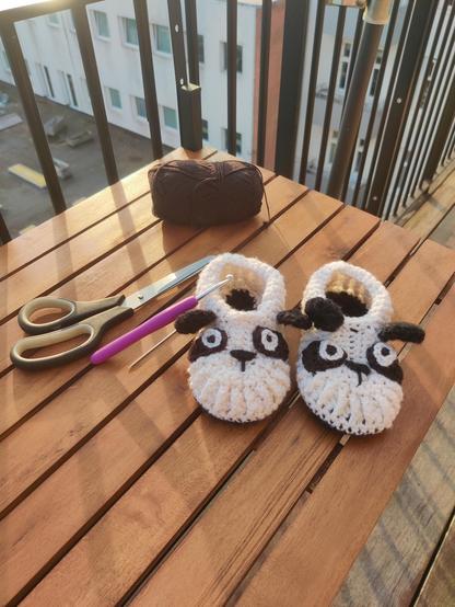A photograph of a crochet project on a balcony table. It's two small shoes on black and white, with eyes, nose and ears to look like cute pandas.

Next to the shoes is a needle and crochet hook, and in the background there's some black yarn 