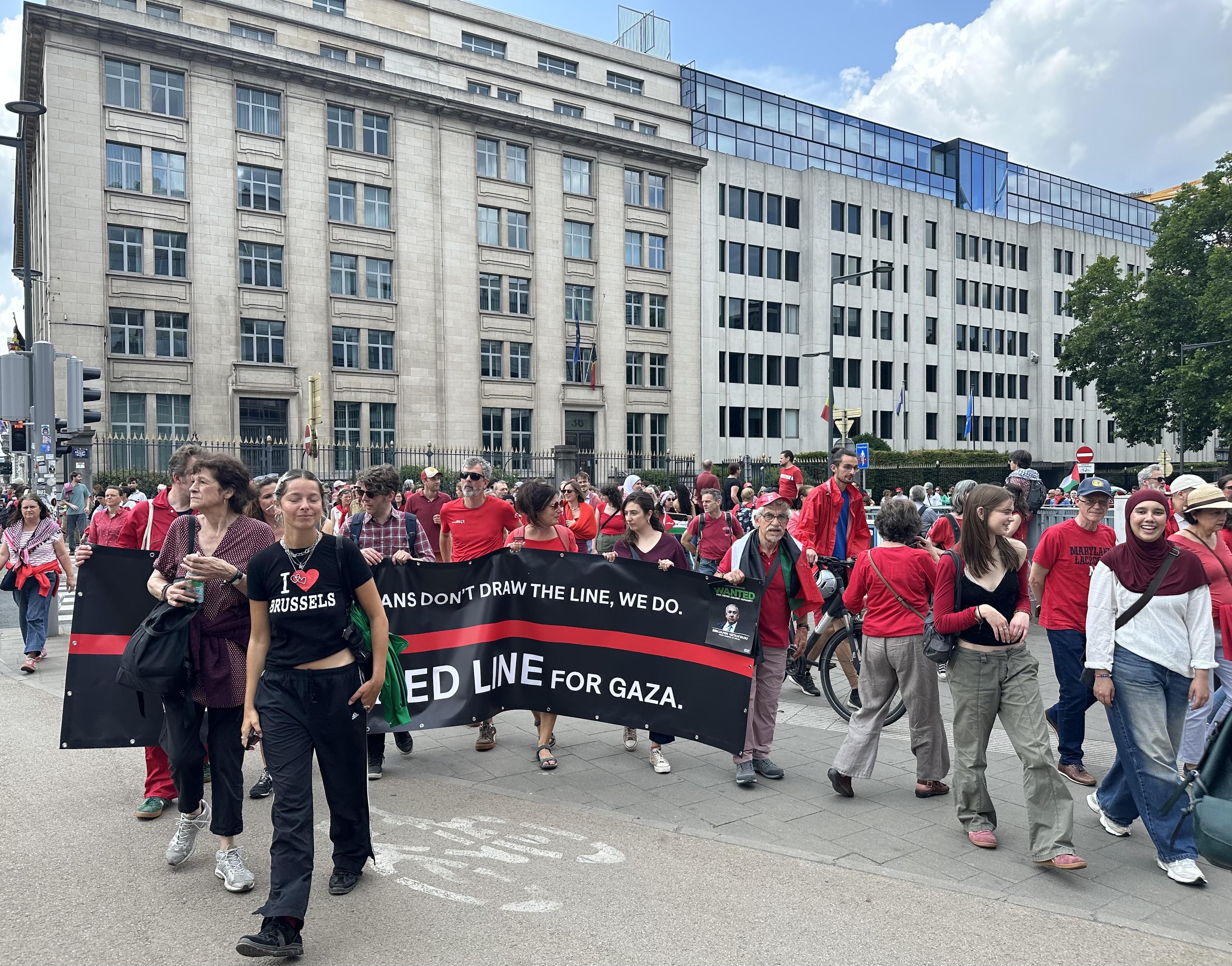 A group of red-clad people carry a banner with the main message of the red-clad demo: “If politicians won’t draw the line, we will”. It’s a black banner with a red horizontal line. Part of the text is blocked by a black clad woman with a I heart Brussels T-shirt 👕