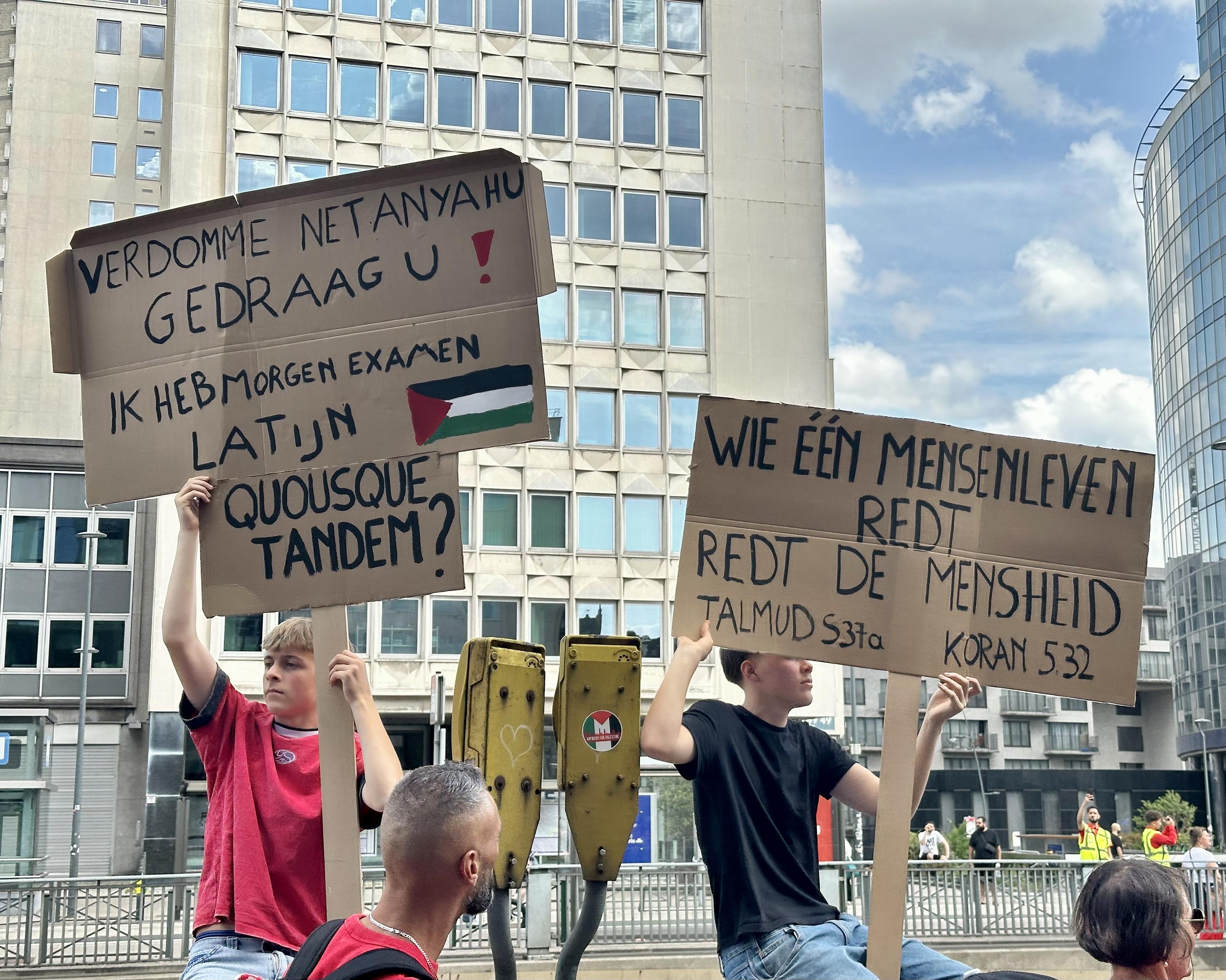 Two Flemish teenage boys are sitting on a fence showing their Dutch-language signs. One says “Damn it Netanyahu, behave yourself! I’ve got Latin exam tomorrow. Quousque Tandem?” The other says “Whoever saves a life saves all humanity. Talmud 537a, Quran 532”