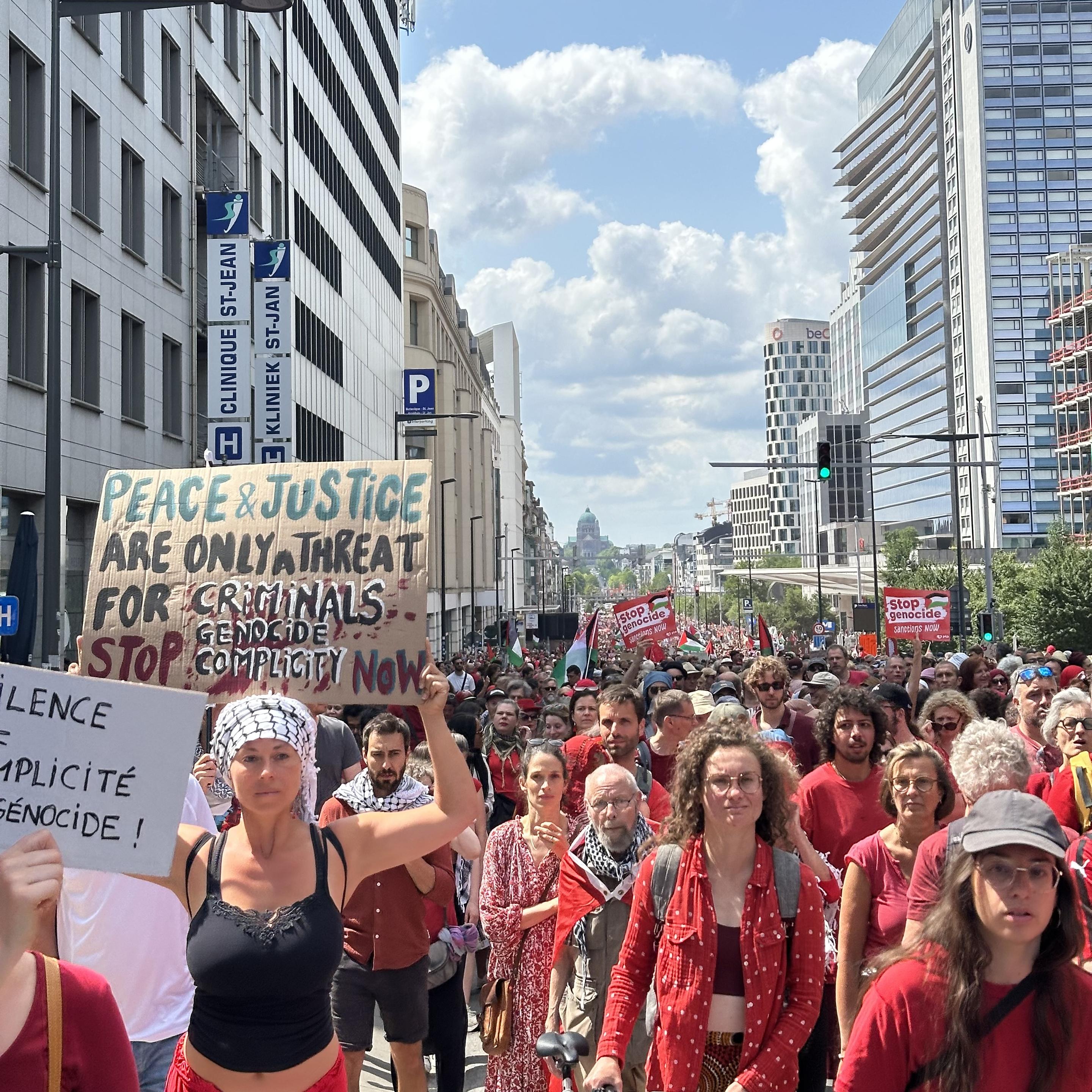 A similar picture as that upthread. Women in the foreground. One carries a sign saying “Peace and Justice are only a threat for criminals. stop genocide / complicity now” ; a French sign to the left repeats that second sentence.