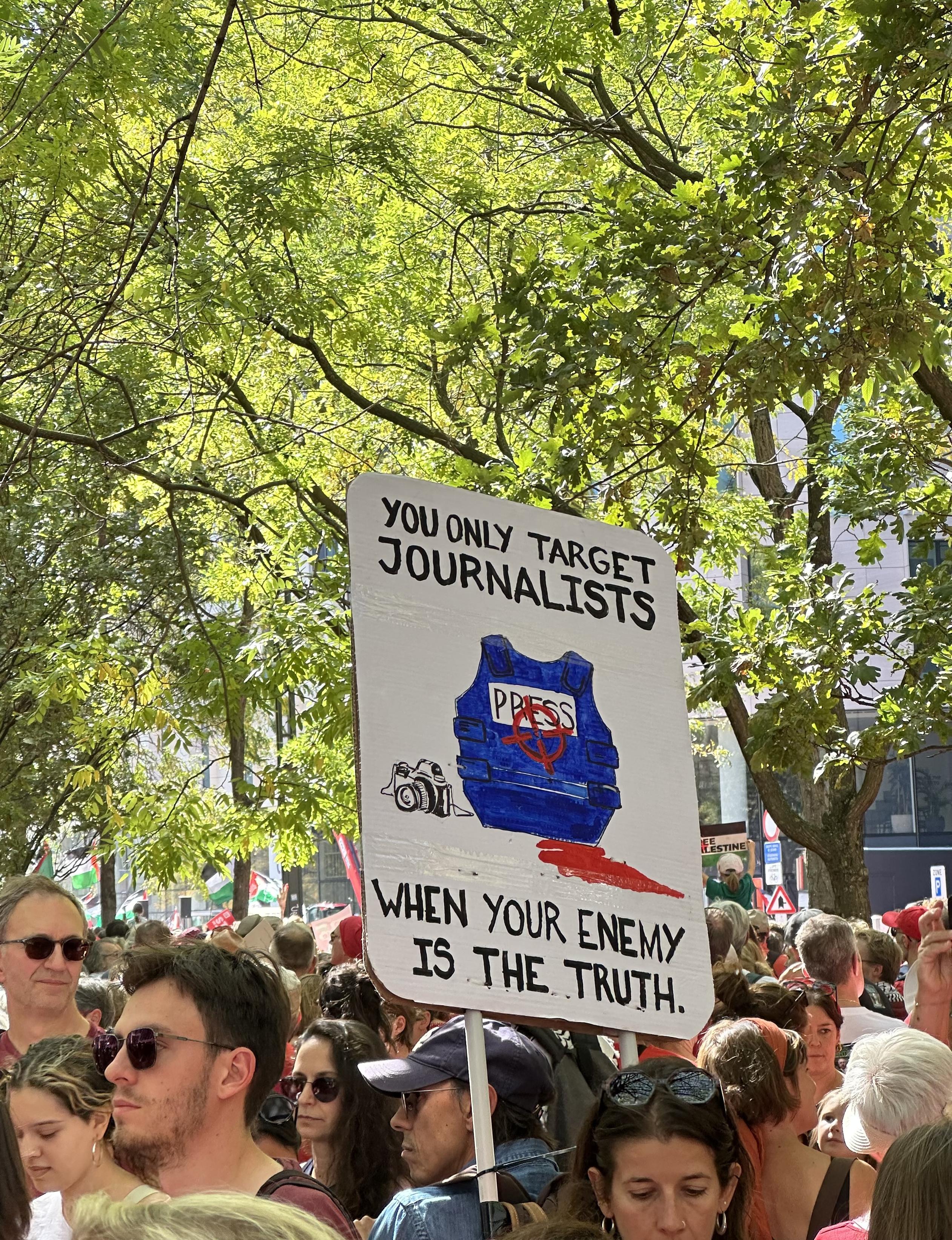 A sign says “you only target journalists when your enemy is the truth.” It shows a bleeding press vest. Trees in the background, people in the foreground