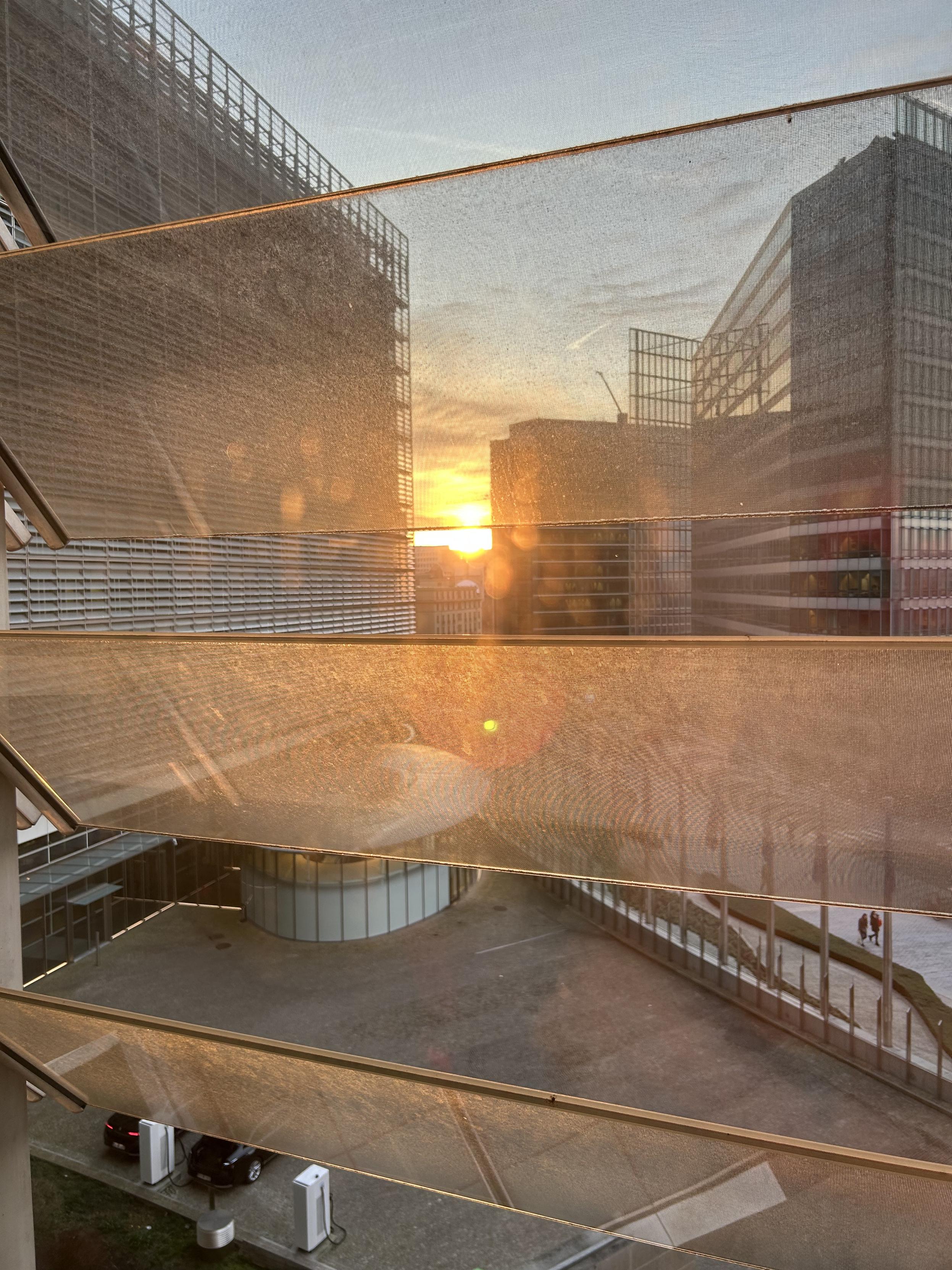 View from an office window onto a setting sun. You can make out the iconic curve of the Berlaymont, the European Commission headquartersโs EU flags line the entrance. Other institutional buildings are around