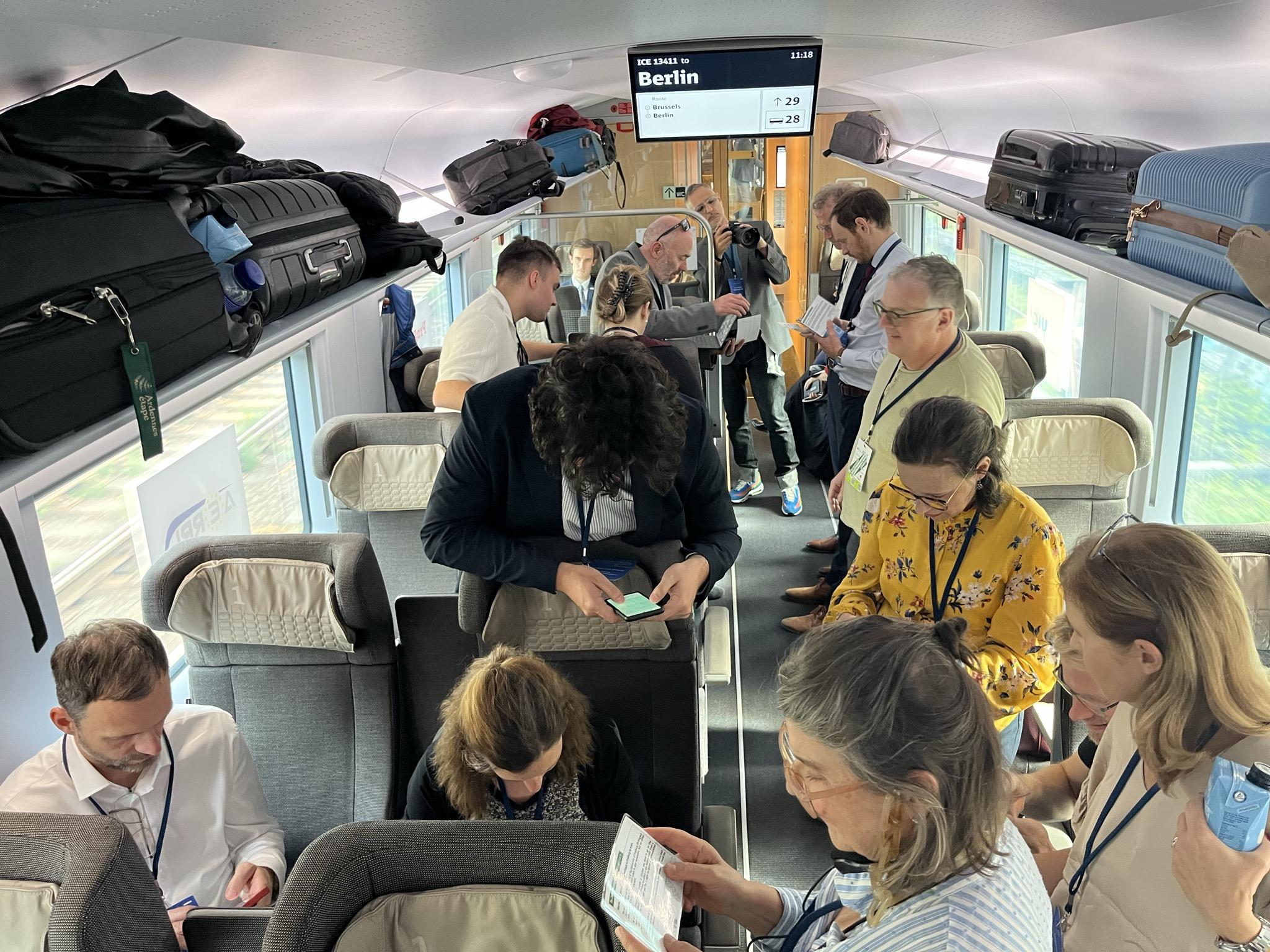 The inside of train with people sitting and standing all energeticaly looking at devices and talking.